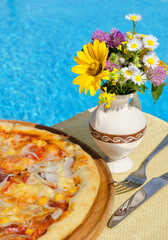 Pizza and vase with wildflowers against the background of a blue pool. Delicious pizza with ham, corn, onions, tomatoes and cheese.