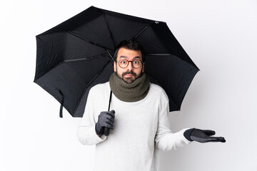 Caucasian handsome man with beard holding an umbrella over isolated white wall having doubts while...