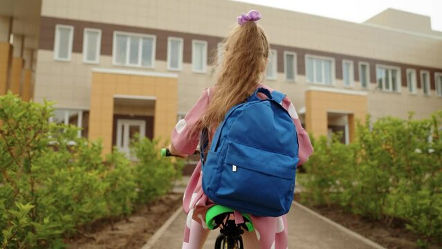 Active cute little girl pupil with backpack driving cycle arriving to elementary school campus back view closeup. Female kid child riding bike on schoolyard enjoy happy childhood sport leisure hobby
