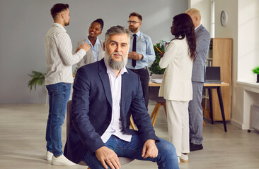 Portrait of mature gray-haired bearded confident business man leader in suit proudly looking at the camera sitting on chair and with a team of company employees talking in background in the office.