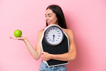 Young caucasian woman isolated on pink background holding a weighing machine while looking an apple