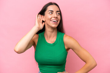 Young caucasian woman isolated on pink background listening to something by putting hand on the ear - obrazy, fototapety, plakaty