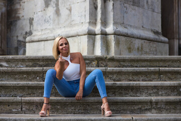 Fototapeta na wymiar Pretty blonde adult woman in jeans and white top sits on the stairs and makes a rude and offensive gesture with her finger. Concept fuck you. The woman is amused.