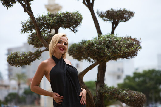 Mature, blonde and beautiful woman wears an elegant black dress and poses for photos next to a garden with an olive tree and flowers. The woman is happy and smiling while doing different expressions.