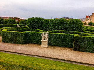 A garden with manicured hedges, two white statues, gravel paths, and a cloudy sky. hedge and sculpture in the park, white sculptures in the park on the background of a green labyrinth of hedgerows