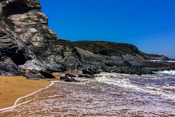 A rocky beach with layered formations, waves crashing, and a cliff under a blue sky with clouds.