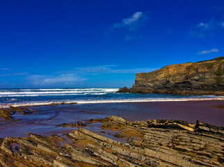 Coastal scene with jagged rocks, turbulent sea waves, and a towering cliff against a clear sky.