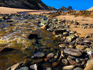 A rocky stream flows into a sandy beach, surrounded by cliffs under a clear sky.