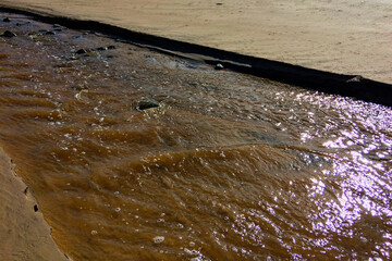 The image captures a muddy stream flowing across a sandy surface, with sunlight reflecting off the wet mud and water.
