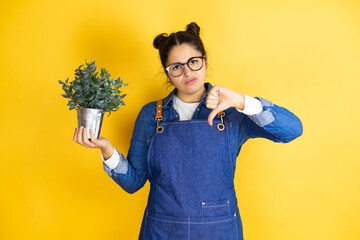 Young caucasian gardener woman holding a plant isolated on yellow background with angry face, negative sign showing dislike with thumb down - obrazy, fototapety, plakaty