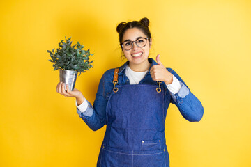 Young caucasian gardener woman holding a plant isolated on yellow background success sign doing...