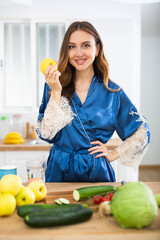 Obraz premium Happy young woman in blue silk peignoir eating apple in home kitchen