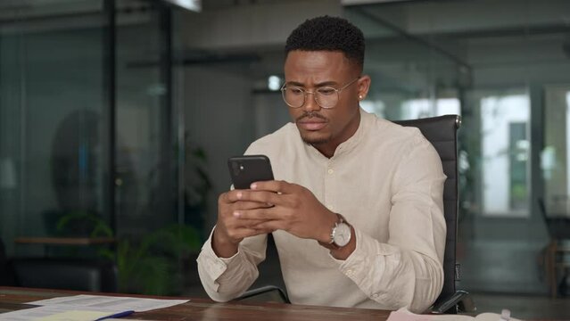 Professional young Black business man holding mobile cellphone looking at cellphone making financial bank payment at work. Busy African American entrepreneur using phone working in office.