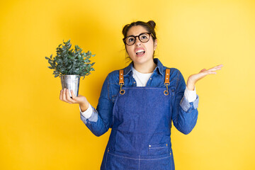 Young caucasian gardener woman holding a plant isolated on yellow background clueless and confused...