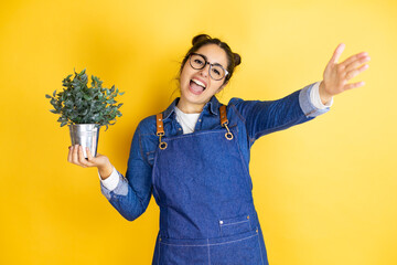Young caucasian gardener woman holding a plant isolated on yellow background looking at the camera smiling with open arms for hug