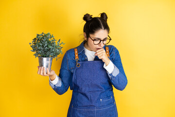 Young caucasian gardener woman holding a plant isolated on yellow background with her hand to her mouth because she's coughing