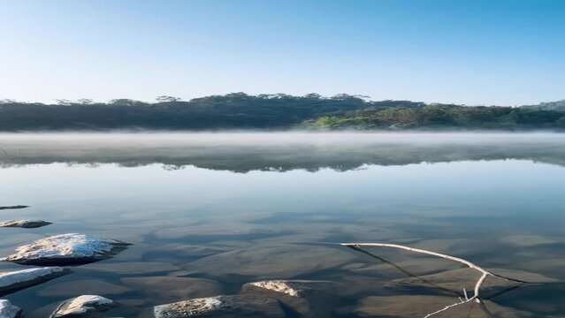Morning mist on serene lake with forest moutain and blue sky background in winter time.