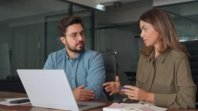 Two happy professional business people, smiling colleagues or company workers talking at meeting using laptop computer discussing online technology project together working in office at workplace.