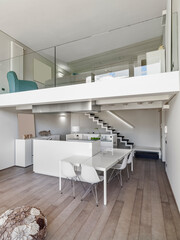 Interior view of a modern kitchen with a mezzanine. In the foreground, at the center of the room, there is a kitchen island. In the background, a staircase leads to the mezzanine. - obrazy, fototapety, plakaty