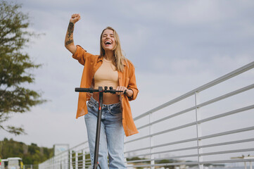 Young smiling overjoyed fun cool calm woman wearing orange shirt casual clothes riding e-scooter do winner gesture walking rest relax in spring city park outdoors on nature. Urban lifestyle concept.