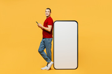 Full body side view young smiling middle eastern man he wearing red t-shirt casual clothes big huge blank screen mobile cell phone with area use smartphone isolated on plain yellow orange background.