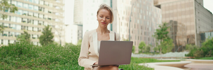 Smiling businesswoman with blond hair wearing beige suit is using pc laptop outside, Panorama