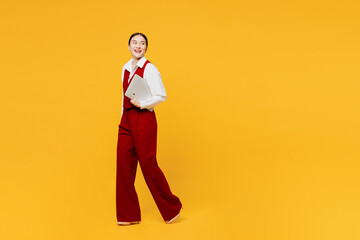 Naklejki  Full body young lawyer employee IT business woman of Asian ethnicity wears red vest shirt work at office walk go hold closed laptop pc computer look aside isolated on plain yellow background studio.