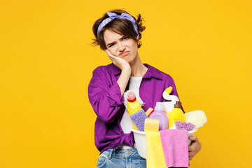 Young sad woman wear purple shirt hold basin with detergent bottles do housework tidy up look camera prop up face look camera isolated on plain yellow background studio portrait. Housekeeping concept.