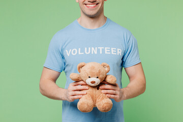 Close up young fun man wear blue t-shirt white title volunteer hold hug teddy bear plush toy isolated on plain pastel light green background. Voluntary free work assistance help charity grace concept