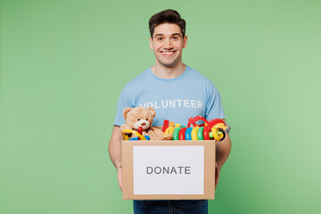 Young happy man wear blue t-shirt title volunteer hold cardboard box with donated toys look camera...