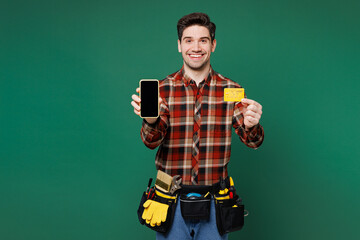Young laborer man wear red shirt hold blank screen mobile cell phone credit bank card shopping online isolated on plain green background. Instruments for renovation apartment room Repair home concept