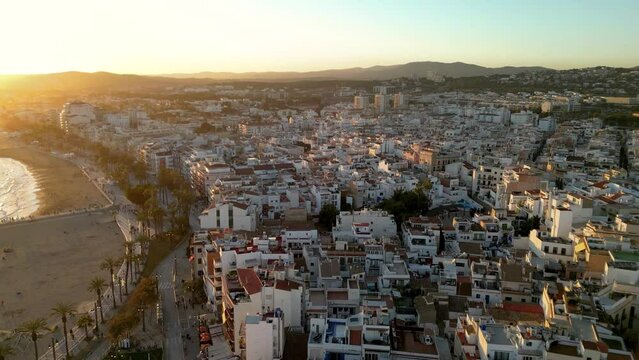 Beautiful orange sunset over Sitges city. Cinematic aerial perspective. Drone going backwards over the Church of Sant Bartolomeu i Santa Tecla. Golden hour colours. Sun reflecting on the buildings.