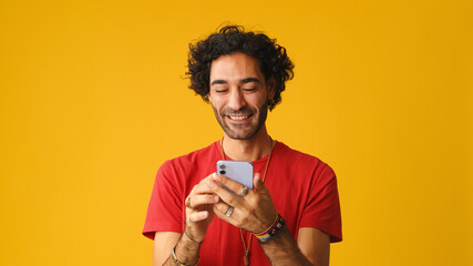 Smiling man with curly hair, dressed in red T-shirt,  using mobile phone, typing massages isolated...