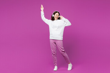 Full body young smiling happy woman wear light hoody casual clothes listen to music in headphones raise up hands have fun isolated on plain purple color background studio portrait. Lifestyle concept.