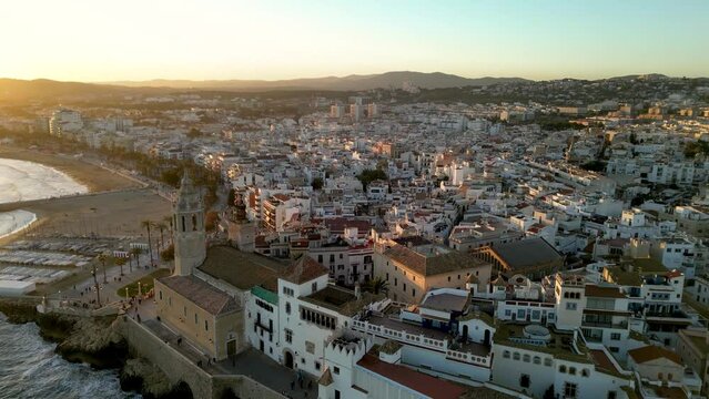 Beautiful orange sunset over Sitges city. Cinematic aerial perspective. Drone going backwards over the Church of Sant Bartolomeu i Santa Tecla. Golden hour colours. Sun reflecting on the buildings.