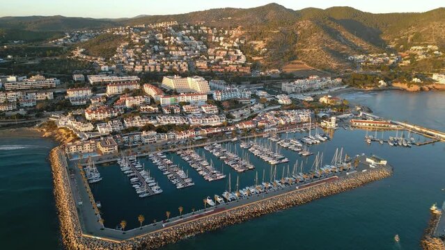 Cinematic aerial view of Sitges Marina at sunset. Drone going forward and camera over the port. Luxury boats and yacht docked in the port. Beautiful warm sunset colours reflecting on the buildings.