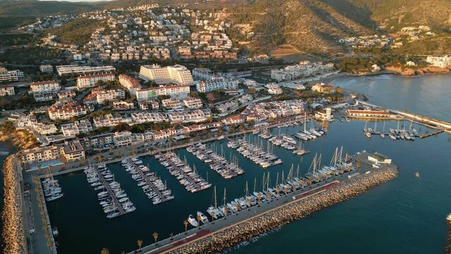 Cinematic aerial view of Sitges Marina at sunset. Drone going forward. Luxury boats and yacht docked in the port. Beautiful warm sunset colours reflecting on the buildings. Famous travel destination