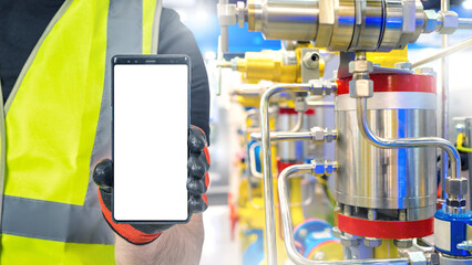 Mobile phone in hands of industrialist. Factory worker holds smartphone. Phone with blank screen. Cellphone near industrial equipment. Person in reflective vest recommends apps. Industrial mock up - obrazy, fototapety, plakaty