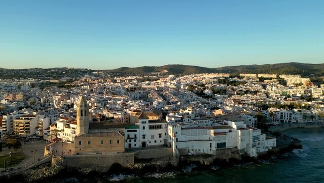 Beautiful orange sunset over Sitges city. Cinematic aerial perspective. Drone rotating around the Church of Sant Bartolomeu i Santa Tecla. Golden hour colours. Sun reflecting on the buildings.
