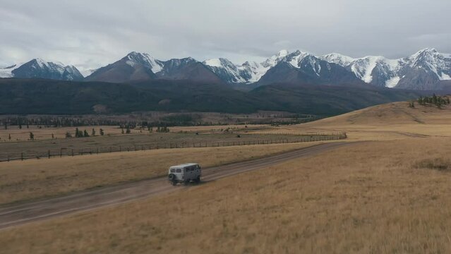 Aerial view gray car microbus driving on rural road steppes. Picturesque mountains snowy caps in wild nature. Road car trip, adventure travel tourism vacations. Behind the fence tractor plows a field