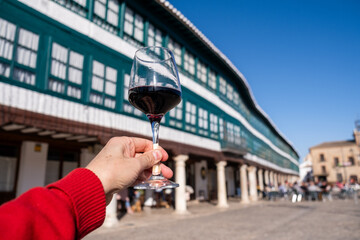 A man's hand holds a glass of wine with the main square of Almagro in the background out of focus on a sunny day.