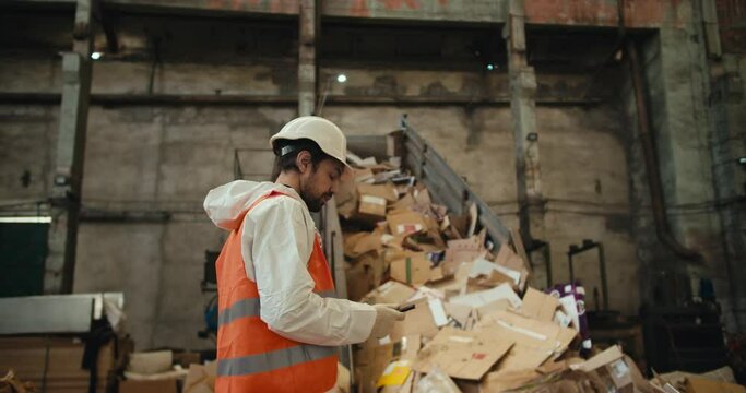 A brunette man with a beard in a white helmet and a white uniform in an orange vest stands near a conveyor belt at a waste paper recycling plant