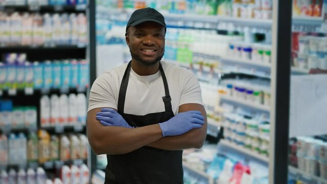 Portrait of a happy man with Black skin in a white T-shirt and a black apron who folded his arms on his chest near display cases with dairy products in a large grocery supermarket