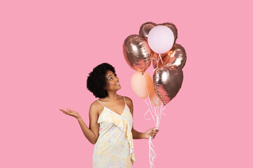 Cheerful African American woman with curly hair, looking up at rose gold and white balloons in her...