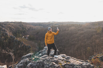Traveller in a yellow jacket standing on the edge of a cliff taking a selfie on his mobile phone at sunset.  Divoka Sarka valley, Prague