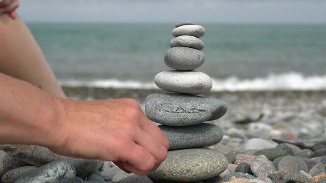 The hands of a woman and a teenager lay out a pyramid of pebbles on the seashore.