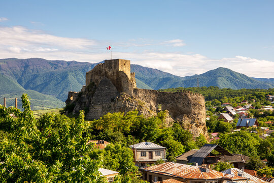 Surami fortress in Georgia, ruins of medieval castle at the top of a hill with Georgian national flag on top, rural architecture around, Likhi Mountain Range in the background.