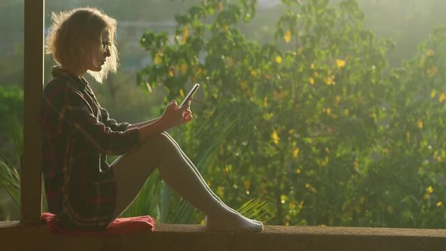 Young caucasian woman sitting on roof terrace at sunset using smartphone for communication. Relaxed female blonde listening music outdoors. Modern technology concept.
