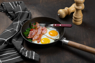 Fried eggs in a frying pan with bacon. Dark background. Salt and pepper shaker on the table.