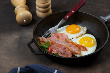 Fried eggs in a frying pan with bacon. Dark background. Salt and pepper shaker on the table.